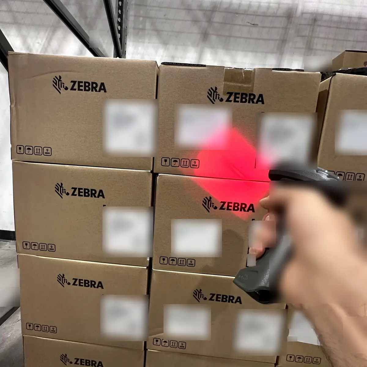 A handheld barcode scanner reading the label on a stacked case of Zebra hardware, its red aiming beam on the carton
