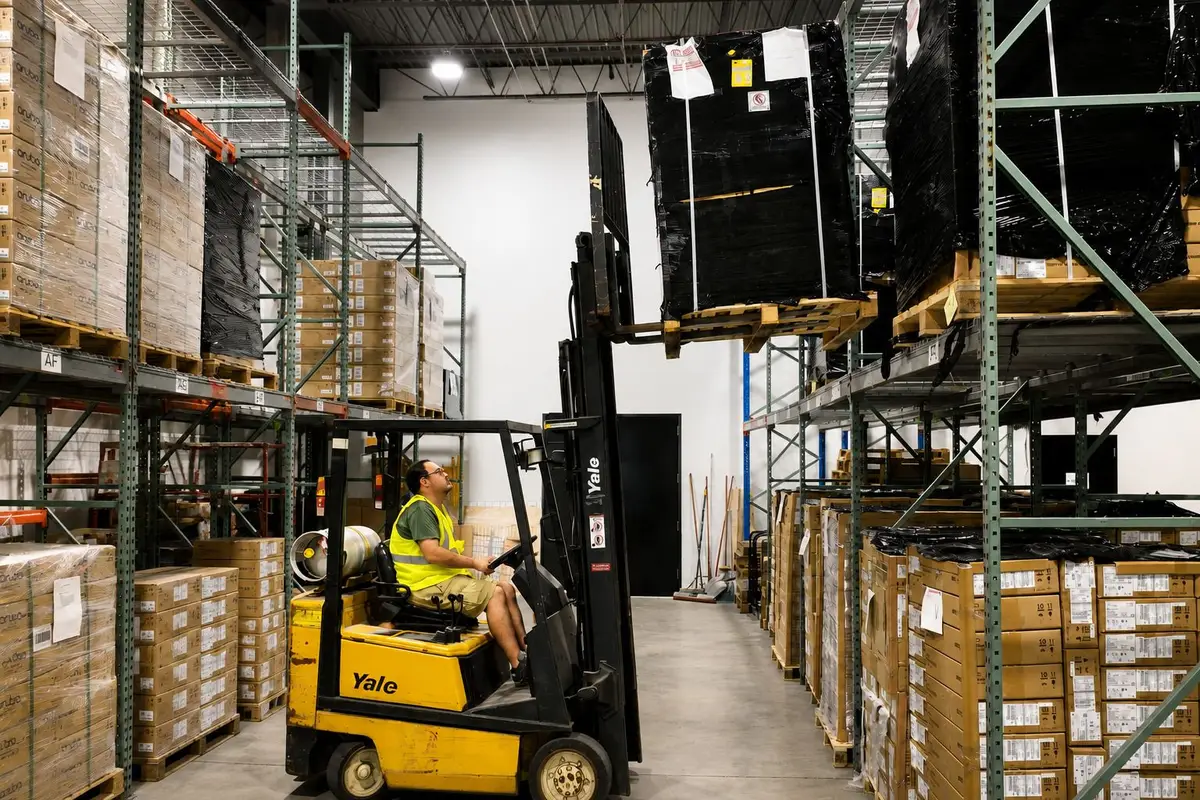 A forklift operator in the 2B Global warehouse raising a shrink-wrapped pallet to the top rack, cartons racked down both sides of the aisle