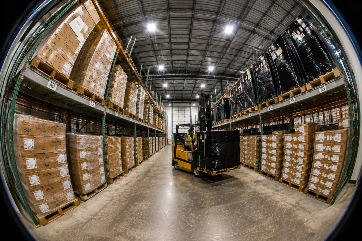 A fisheye view down a racked aisle of the 2B Global warehouse, a forklift carrying a shrink-wrapped pallet between pallets stacked to the roof on both sides