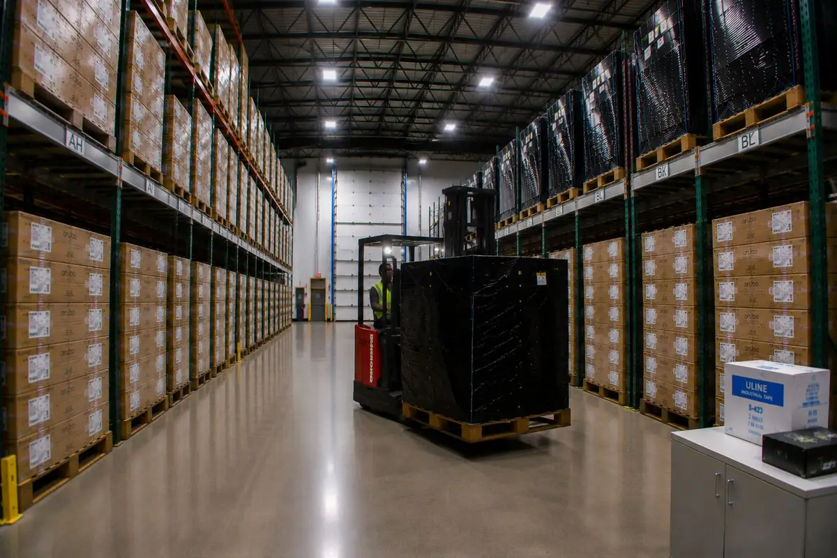 A racked aisle in the 2B Global warehouse, both sides stacked floor to ceiling with cartons, an order picker moving a wrapped pallet down the middle