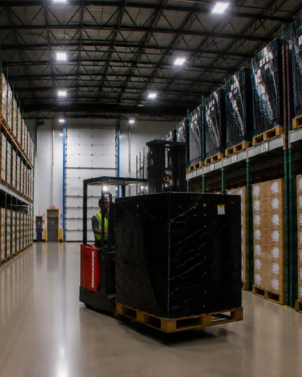 A reach-truck operator carrying a wrapped pallet down the racked aisle of the 2B Global warehouse, cartons stacked to the roof on both sides and the dock doors at the far end